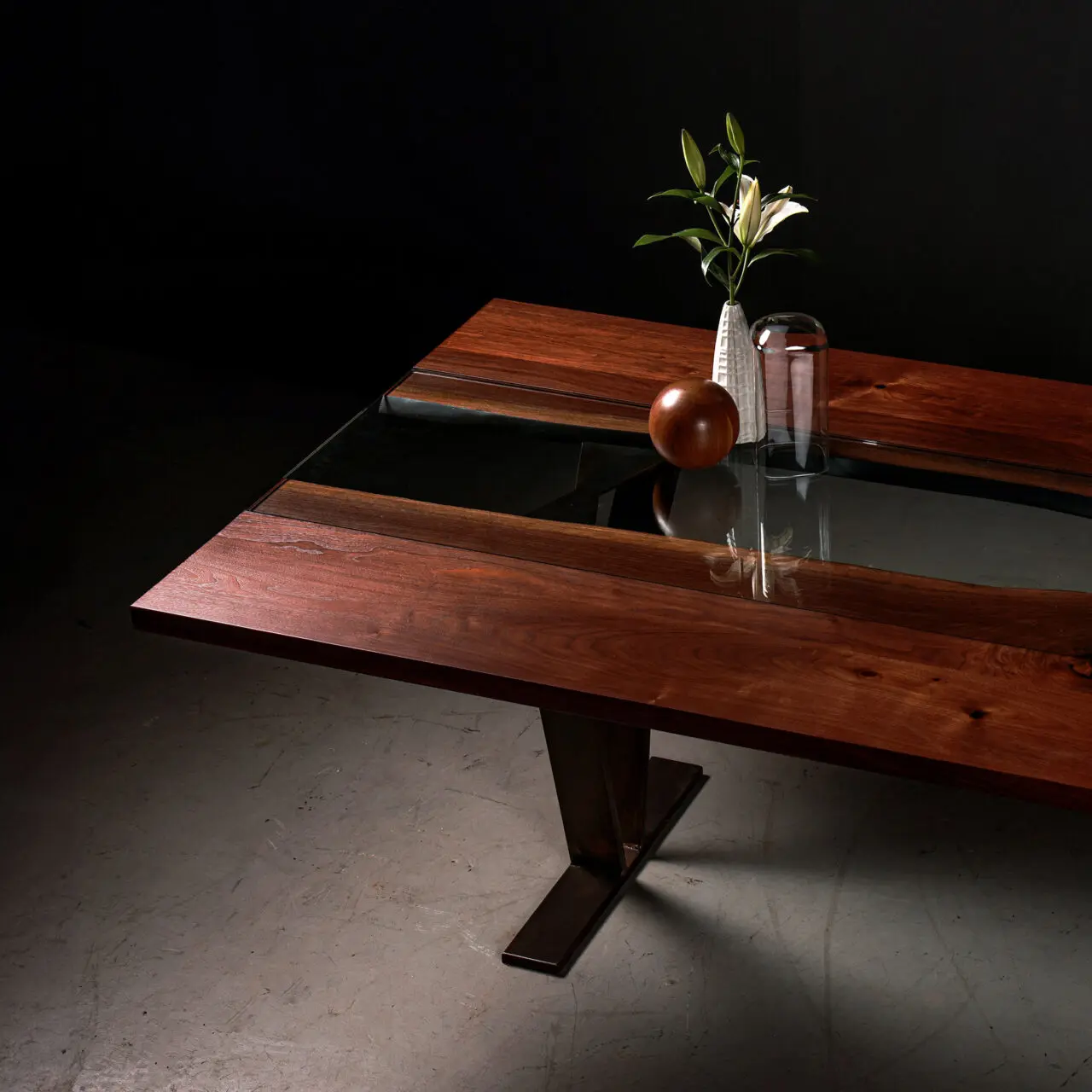 Colorado, a modern dining table by SENTIENT Furniture with a rich, dark wood surface and a single leg base. A glass vase with a green plant and a wooden spherical object are placed on top, reflecting on the glass panel that runs through the center of the table.