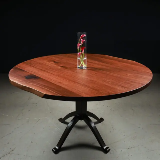 Flow, a pedestal round dining table by SENTIENT Furniture with a beautiful reddish wood top and a black metal base. The table is adorned with a tall glass vase containing red and green flowers, set against a black backdrop for a dramatic effect.