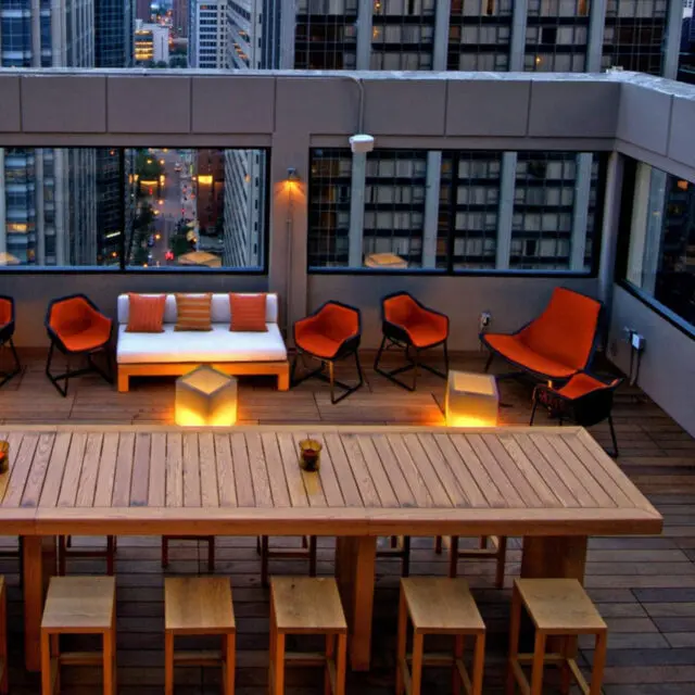 A modern rooftop terrace featuring SENTIENT custom outdoor furniture, including a large wooden dining table, stools, and lounge seating with orange cushions. The design offers a perfect blend of comfort and urban sophistication.