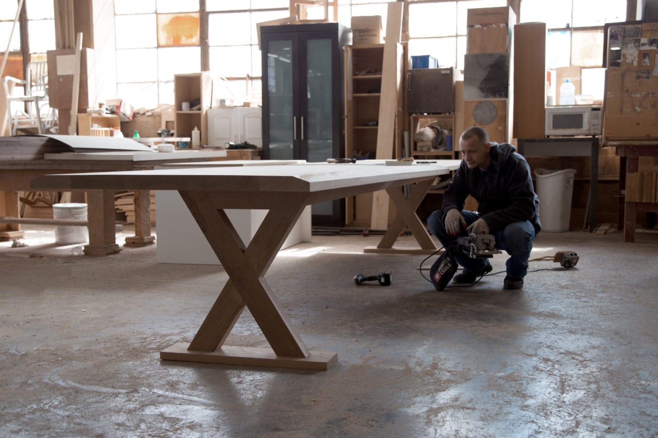 in a workroom a SENTIENT custom maple wood table with x shaped base and a man crouched surveying his work
