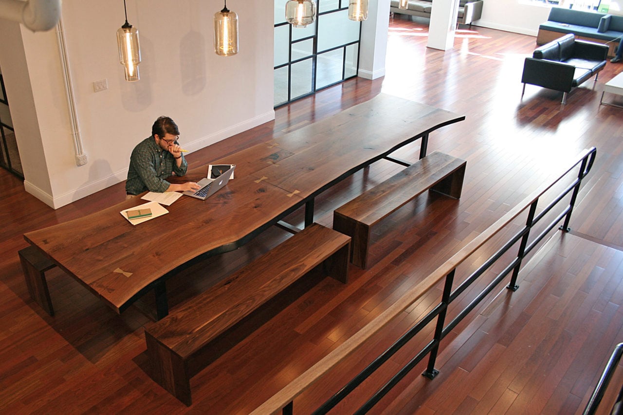 A SENTIENT contemporary live edge custom walnut table with benches and a man sitting and working in an open office setting 