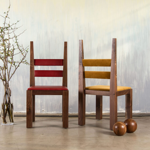 A pair of DC1 unique wooden chairs by SENTIENT Furniture, one with a red seat and the other with a mustard yellow seat. They exhibit a modern, clean-lined design and are staged against a backdrop of a white painted wall and a wooden floor with decorative branches and wooden spheres nearby.