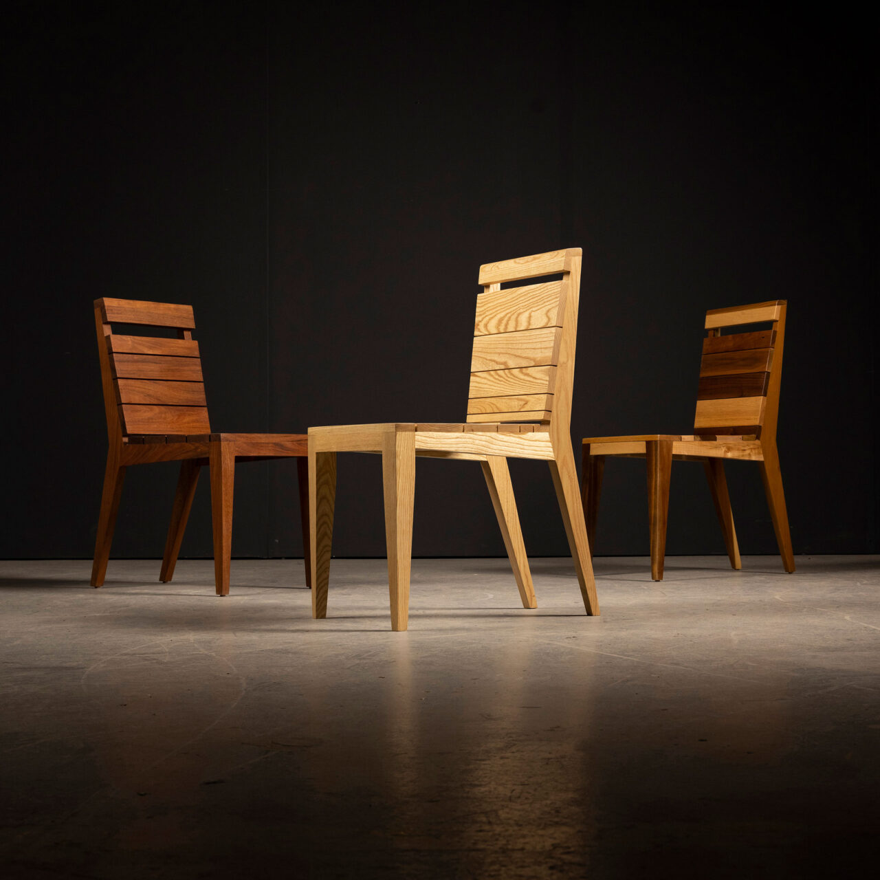 A trio of wooden Angles Unique Chairs by SENTIENT Furniture, each featuring a different grain and color of wood, is positioned on a dark floor against a black backdrop, emphasizing the unique woodwork and craftsmanship.