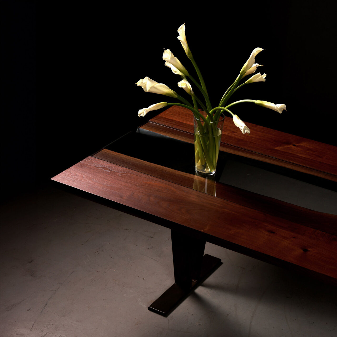 The Colorado dining Table by SENTIENT Furniture, shown in a dimly lit room, is a sleek, dark wooden table with a reflective surface, adorned by a vase with white flowers, creating an elegant and serene dining atmosphere.