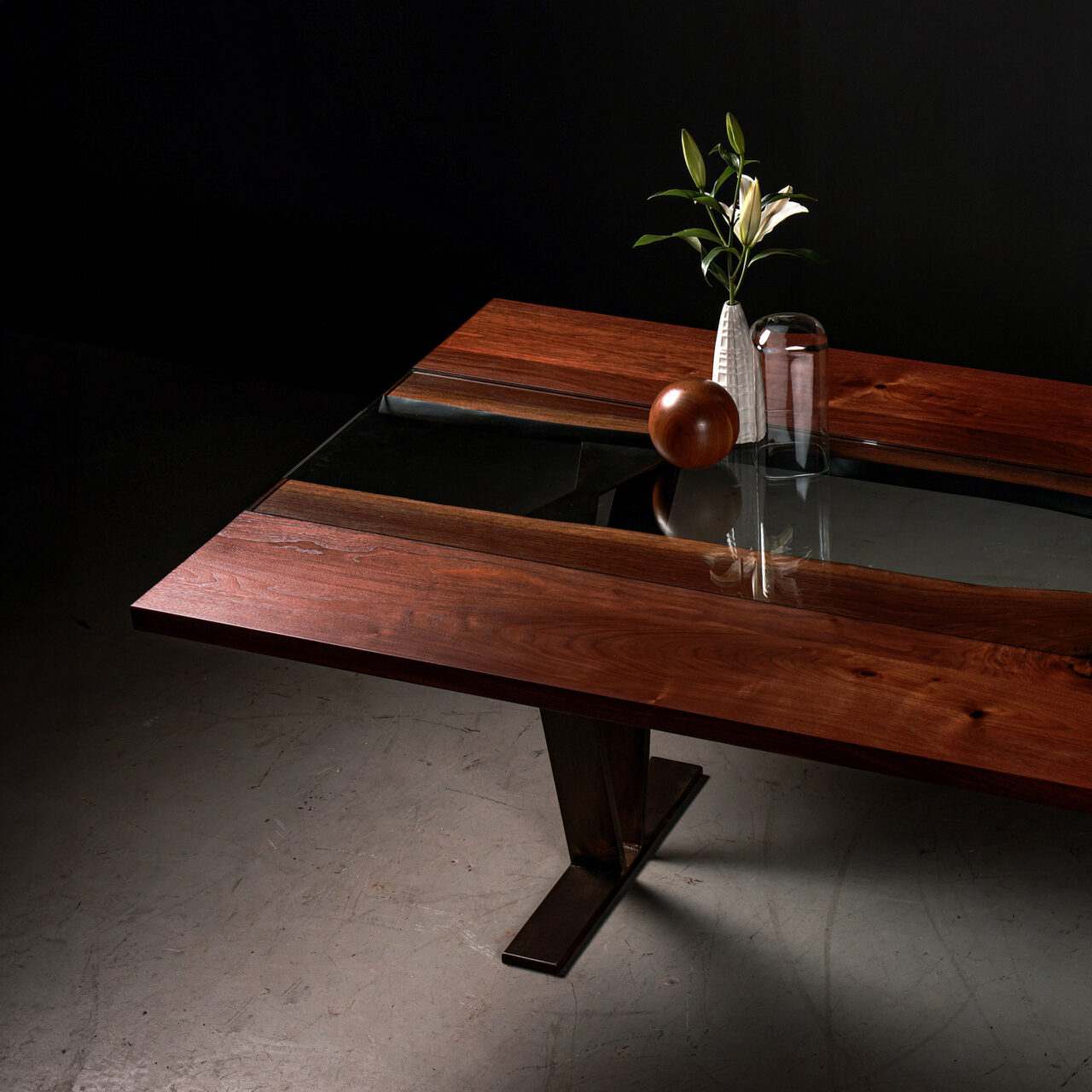 Colorado, a modern dining table by SENTIENT Furniture with a rich, dark wood surface and a single leg base. A glass vase with a green plant and a wooden spherical object are placed on top, reflecting on the glass panel that runs through the center of the table.