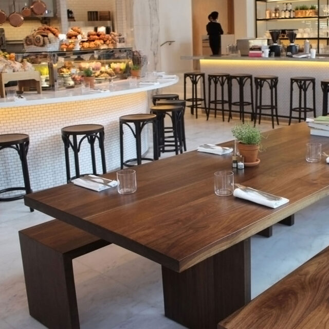 A long, communal dining table by SENTIENT Furniture situated in a bright café setting. The dark wood table is accompanied by simple black stools and is set against a white tiled bar with food items on display.