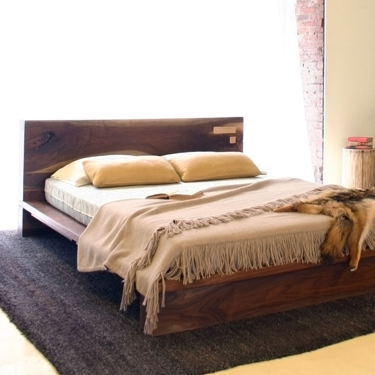 A cozy bedroom setting featuring the SENTIENT Liffey bed, a king-size platform bed made of dark walnut with a large headboard and a tan throw blanket, placed on a dark rug.