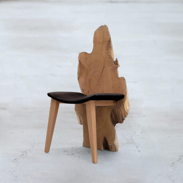 A unique wooden chair by SENTIENT Furniture, featuring an organic, live edge, asymmetrical backrest that mimics the natural form of a tree. The seat is upholstered with black fabric, providing a soft contrast to the solid oak construction. The chair stands on simple, sturdy legs, blending artisanal craftsmanship with a touch of modern design.