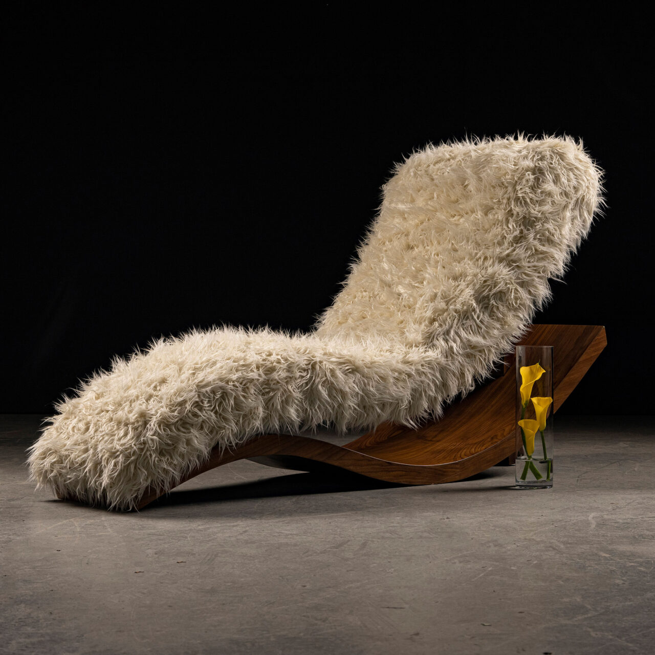 The luxury lounge chair, 'Wave Lounge II' by SENTIENT Furniture, a curvaceous wooden lounge chair covered in fluffy white fur, accompanied by a small yellow sculpture in a clear vase.