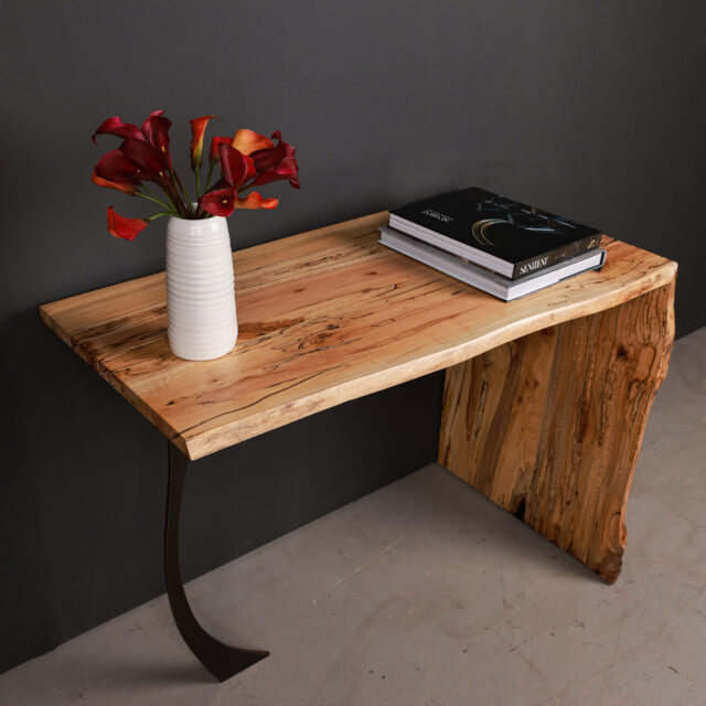 A side view of the Arcady Waterfall Desk crafted in spalted maple, featuring a sculptural design with natural grain patterns and a curved leg accent.