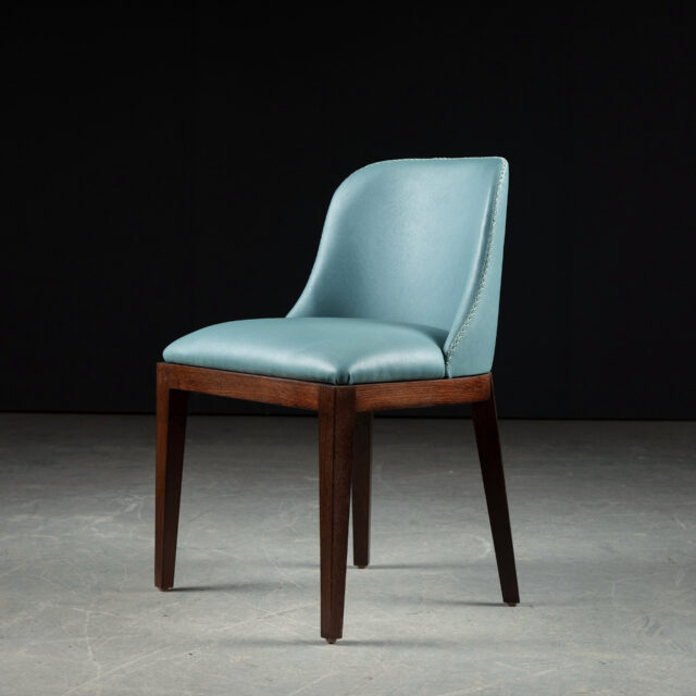 A Lurra dining chair upholstered in muted sage green leather paired with a minimalist accent table featuring a natural wood finish and a vase of yellow calla lilies.